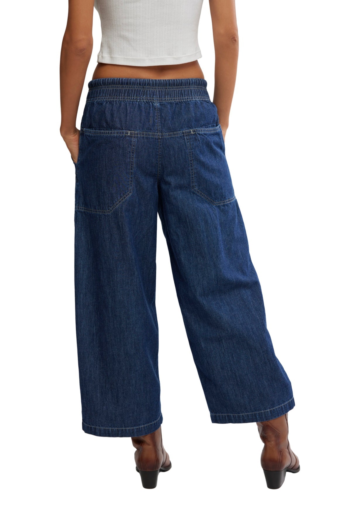Someone wearing dark blue Free People Easy Peasy Pull On Jeans and a white sleeveless top, standing with hands in pockets, back to camera.