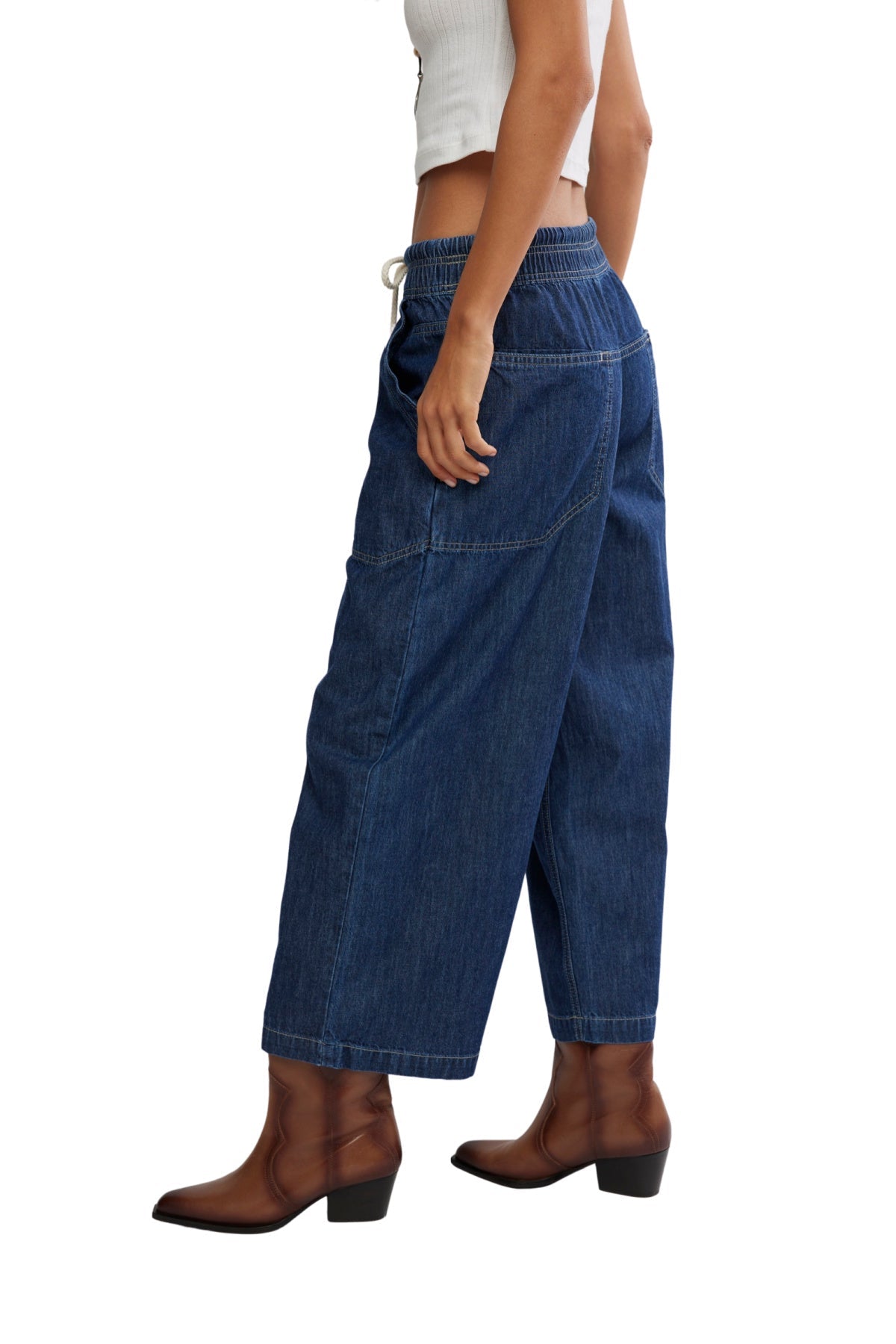 Person in Free People Dark Blue Easy Peasy Pull On Jeans, a white sleeveless top, and brown ankle boots, standing sideways.