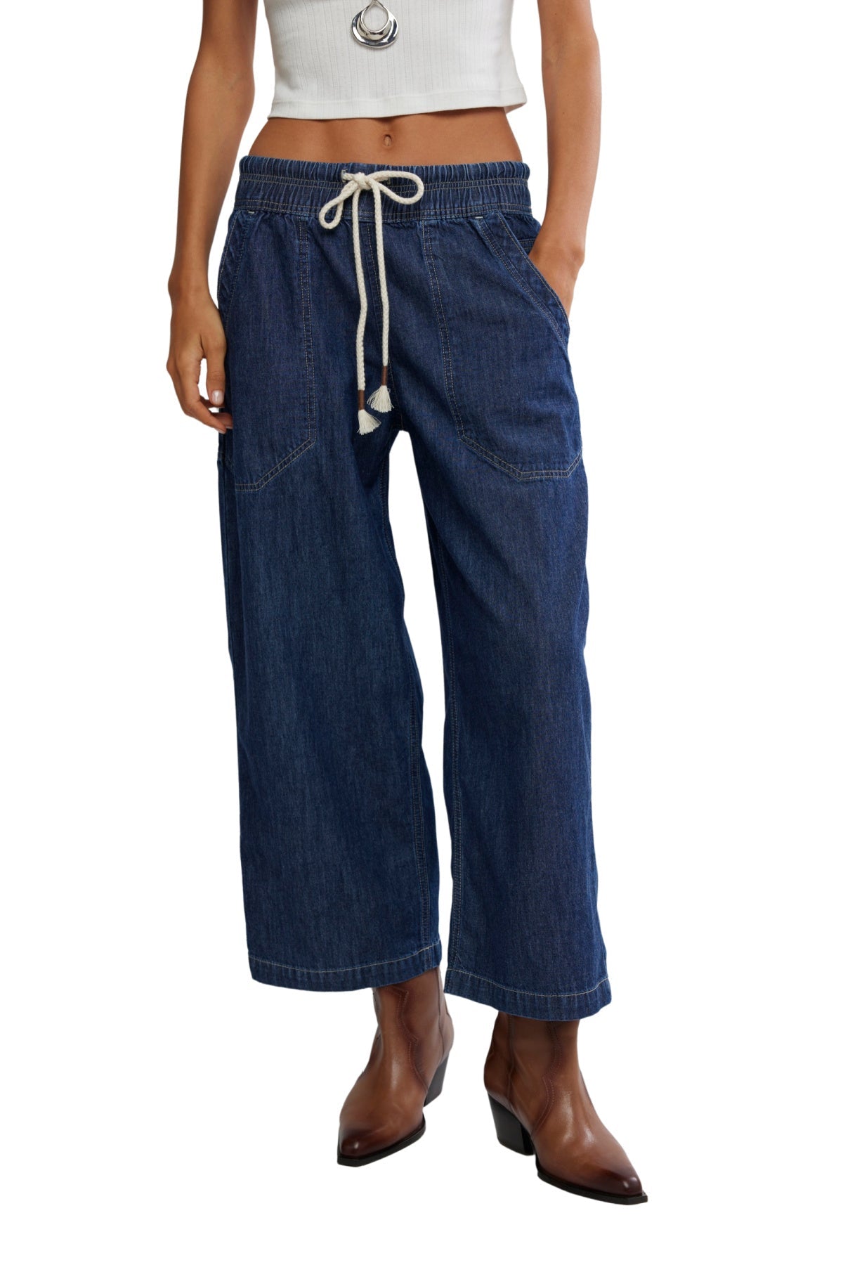 Person wearing a white cropped top, Free People Dark Blue Easy Peasy Pull On Jeans, and brown heeled boots, hands in pockets.