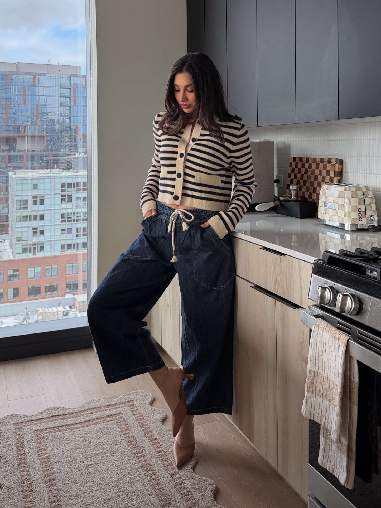A woman in a striped sweater and Free People Dark Blue Easy Peasy Pull On Jeans leans against a modern kitchen counter by a city-view window.