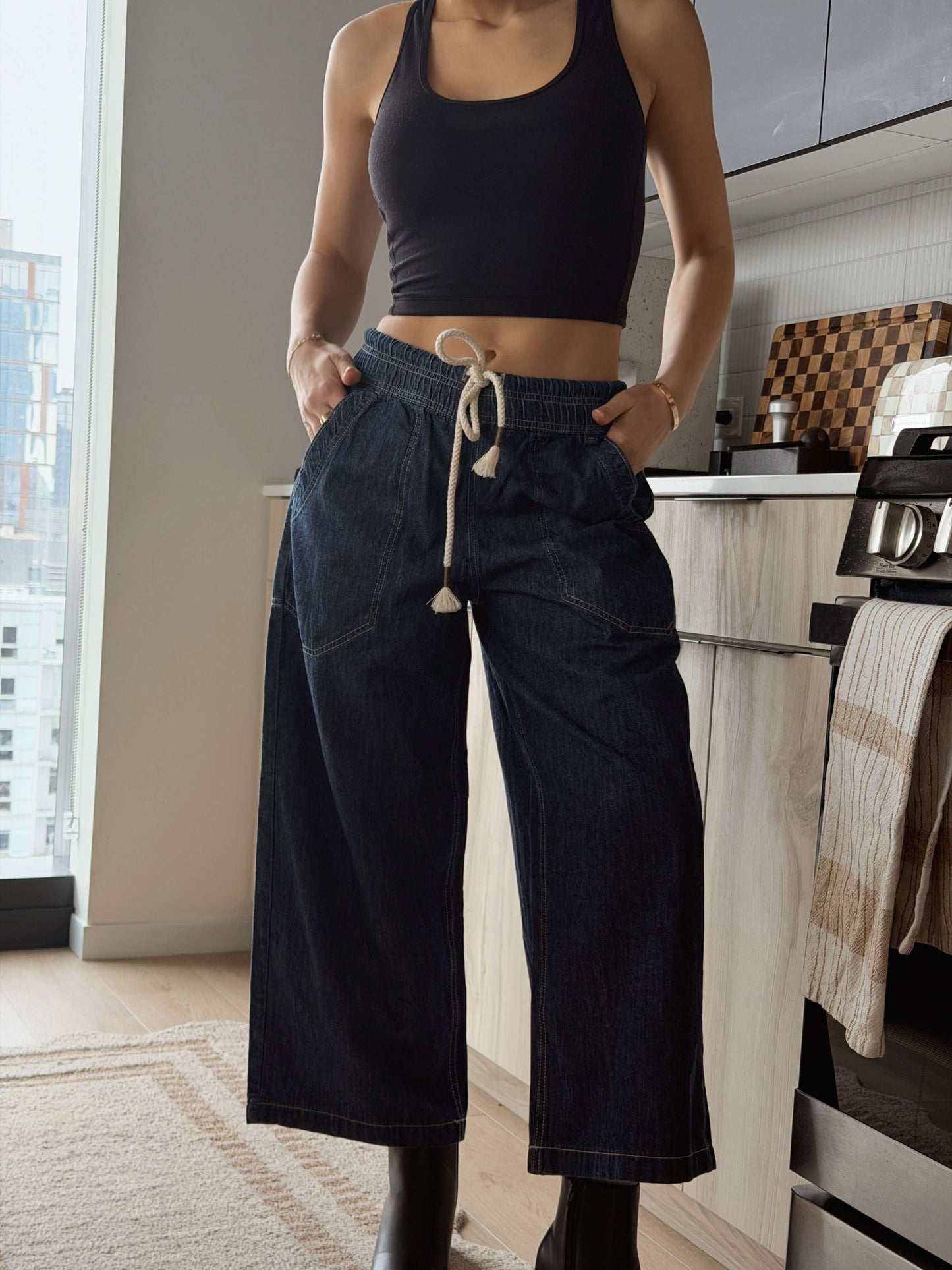 A woman in a black tank top and wide-leg Free People Dark Blue Easy Peasy Pull On Jeans stands in a modern kitchen.