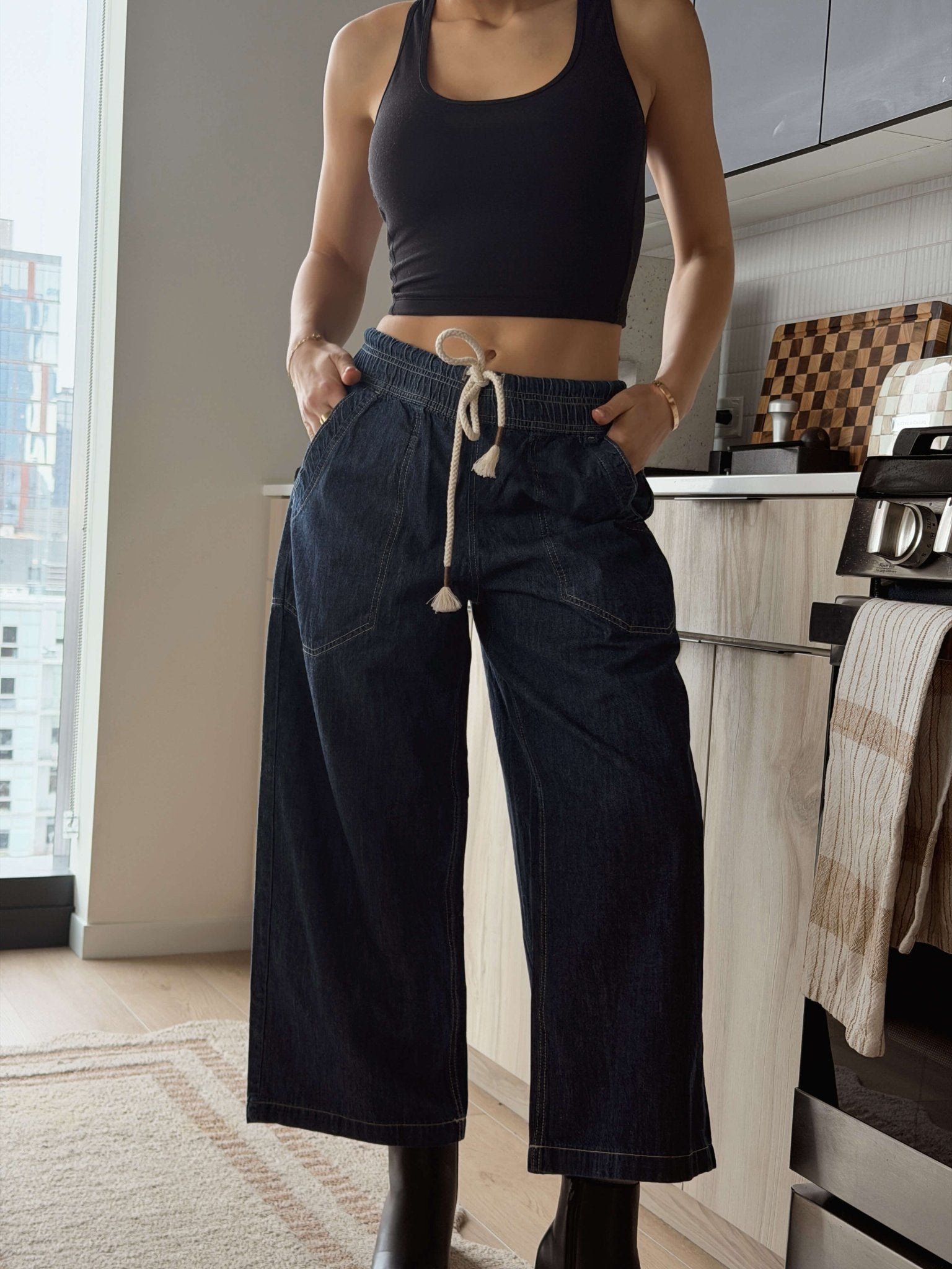 A woman in a black tank top and wide-leg Free People Dark Blue Easy Peasy Pull On Jeans stands in a modern kitchen.