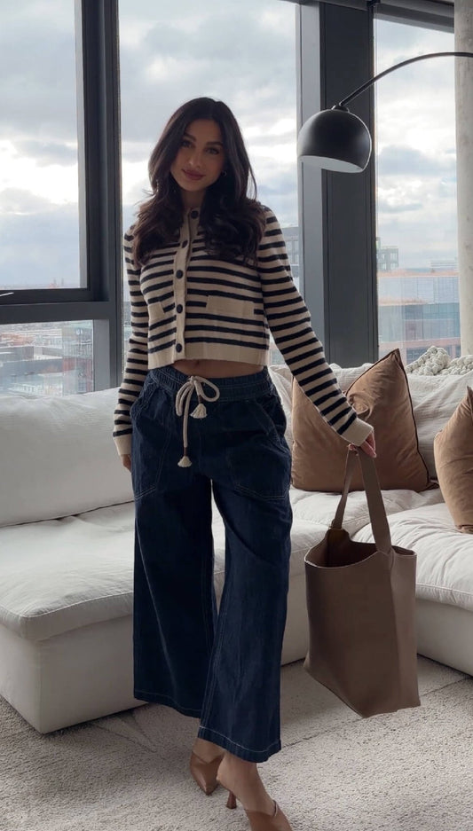 Woman stands in a modern living room, wearing a striped cardigan, Free People Dark Blue Easy Peasy Wide Leg Pants, and holding a brown tote.