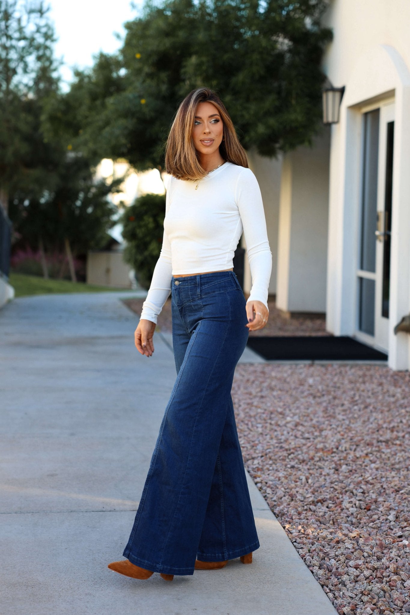 A woman in a white long-sleeve top and Free People Dark Wash Libre Wide Leg Jeans stands outside near a building and trees.