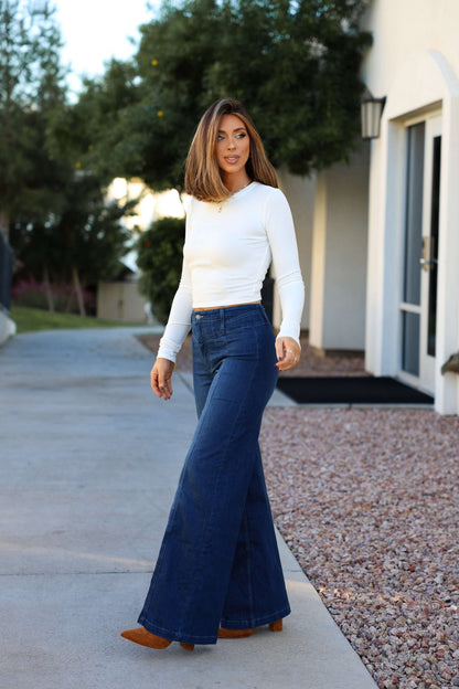 A woman in a white long-sleeve top and Free People Dark Wash Libre Wide Leg Jeans stands outside near a building and trees.