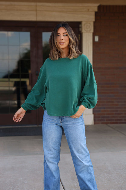 Woman with shoulder-length hair wears a Free People Forest Green Total Eclipse Tee and blue jeans, standing outside near glass doors.
