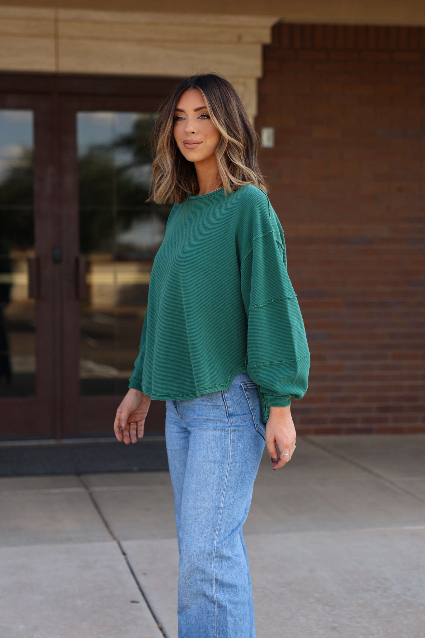 Woman with shoulder-length wavy hair in a Free People Forest Green Total Eclipse Tee and blue jeans, outside a brick building.