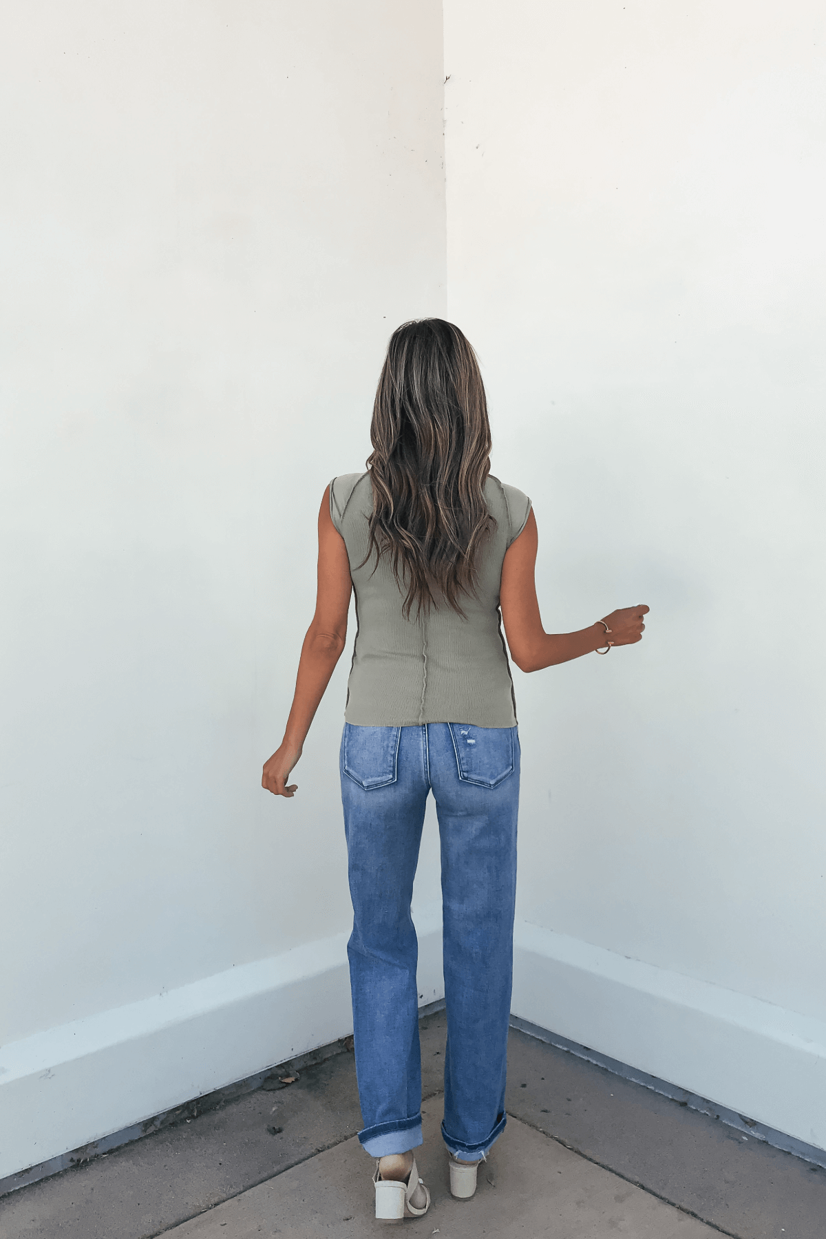 A woman with long wavy hair faces a white wall, wearing the Free People Green Ever Had Tee, blue jeans, and sandals.