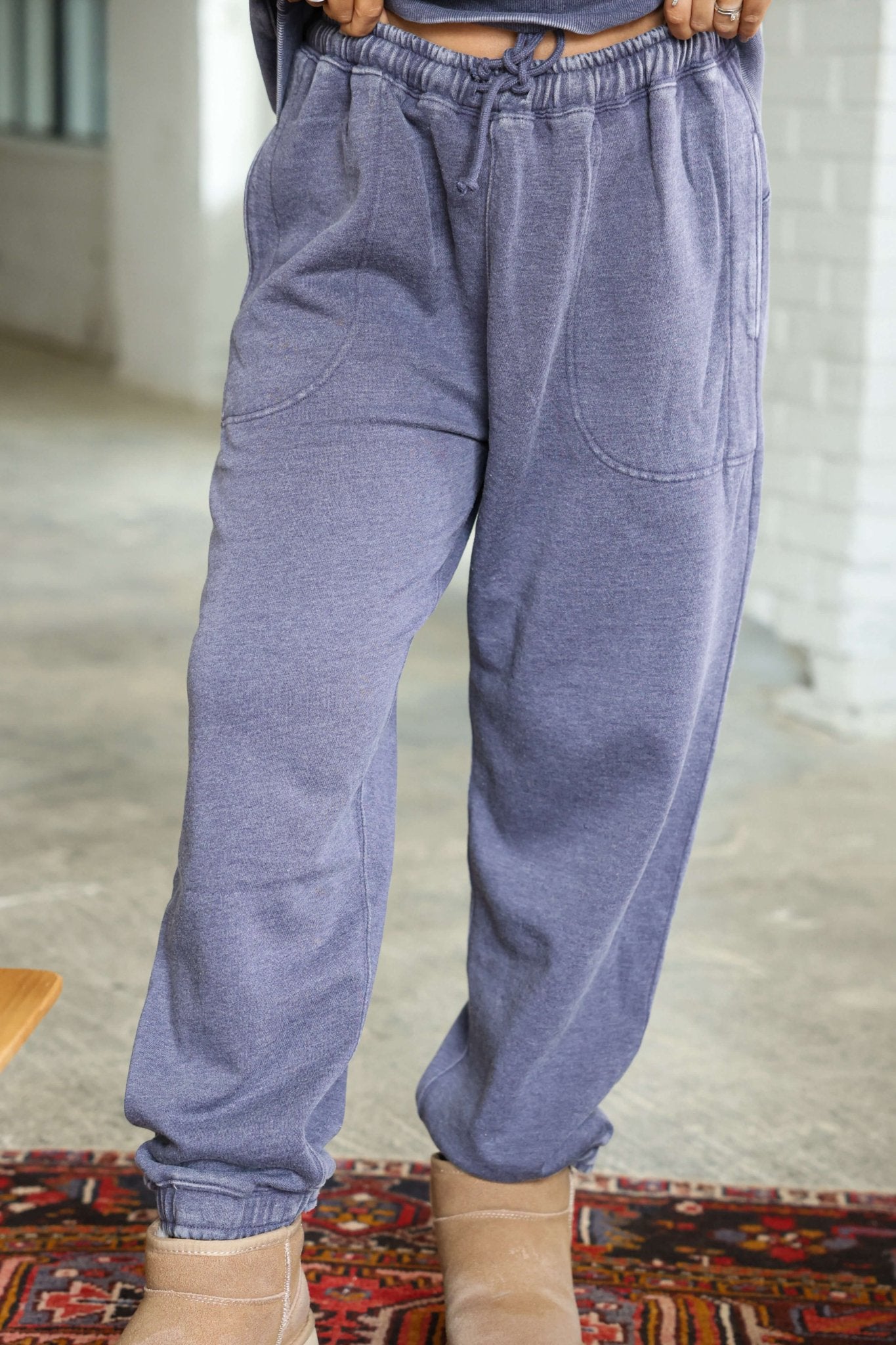 Person wearing loose-fitting grey Free People Slow It Down Joggers and tan slippers, standing indoors on a patterned rug.