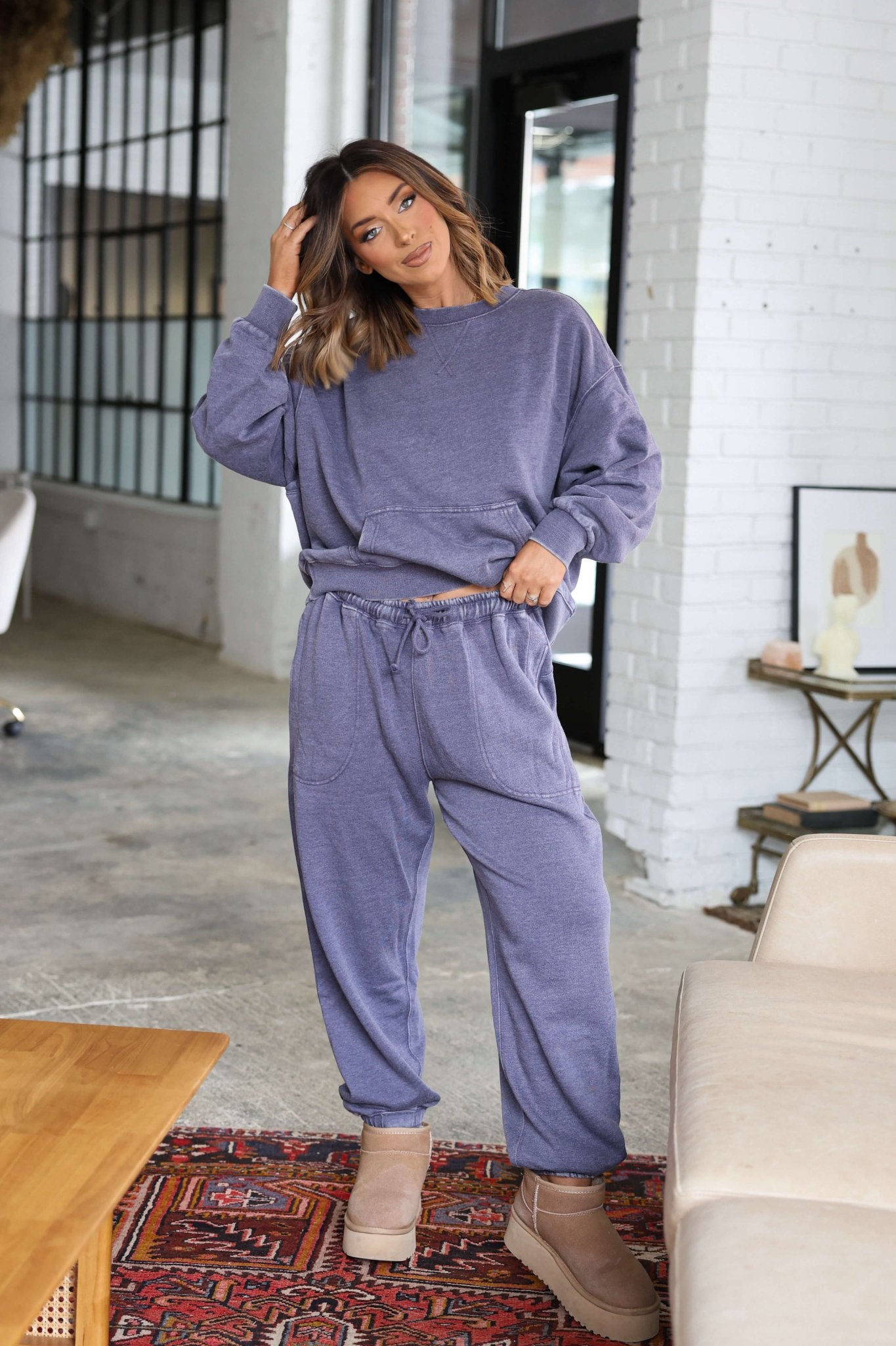 Woman in Free People Grey Slow It Down Joggers and beige boots poses indoors by a couch, touching her hair.