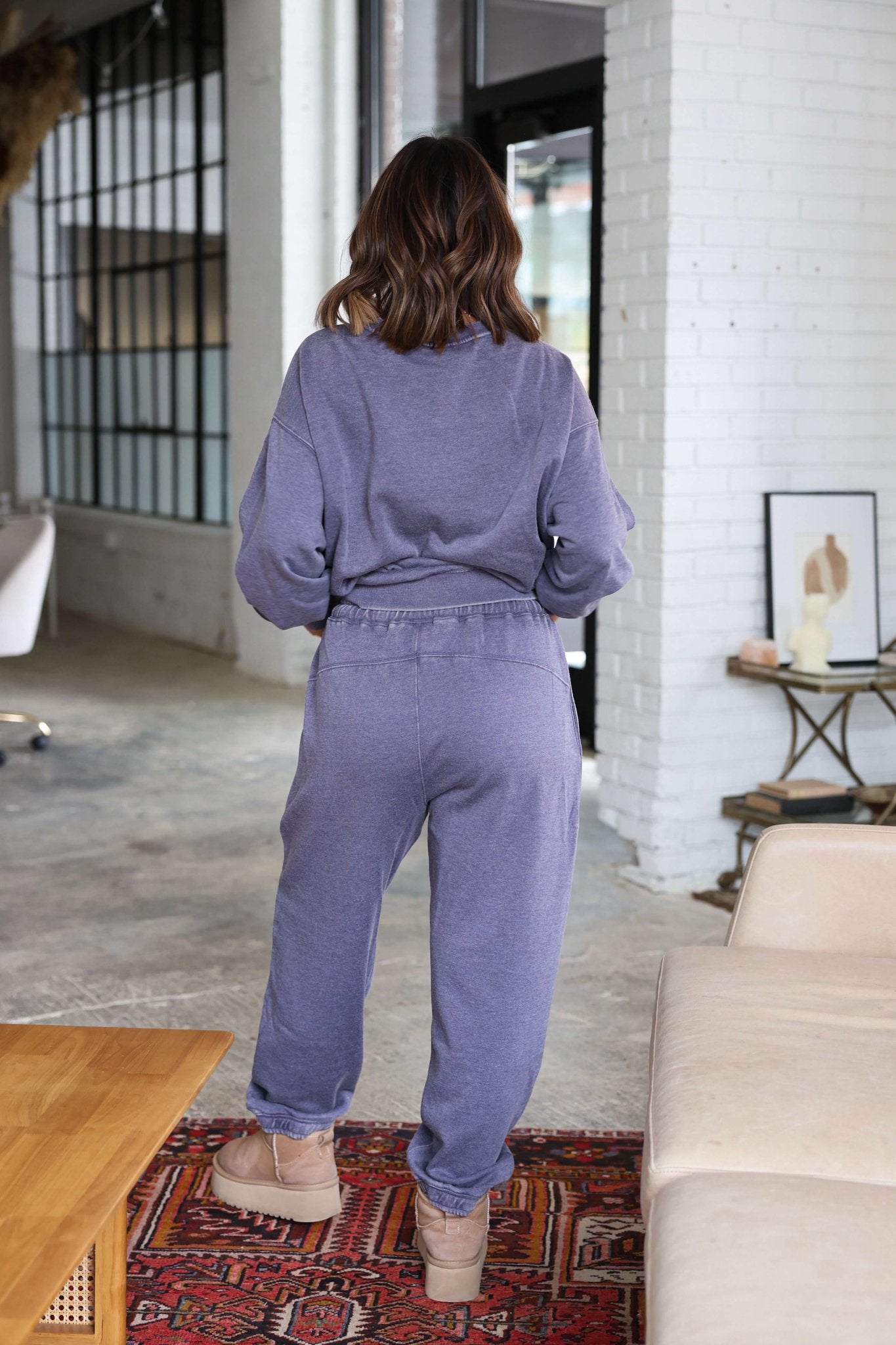 A woman in Free People Grey Slow It Down Joggers and beige boots stands indoors with her back to the camera.