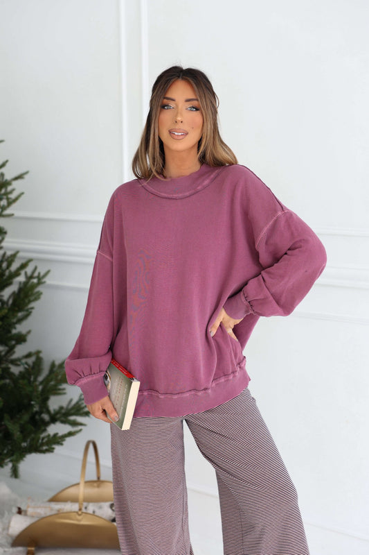 Woman in a loose Free People Iris Orchid Palm Springs Pullover holds a book indoors near a green plant and tan bag.