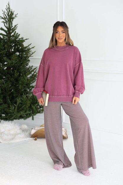 Woman stands indoors by a Christmas tree in a loose Free People Iris Orchid Palm Springs Pullover, holding a book.