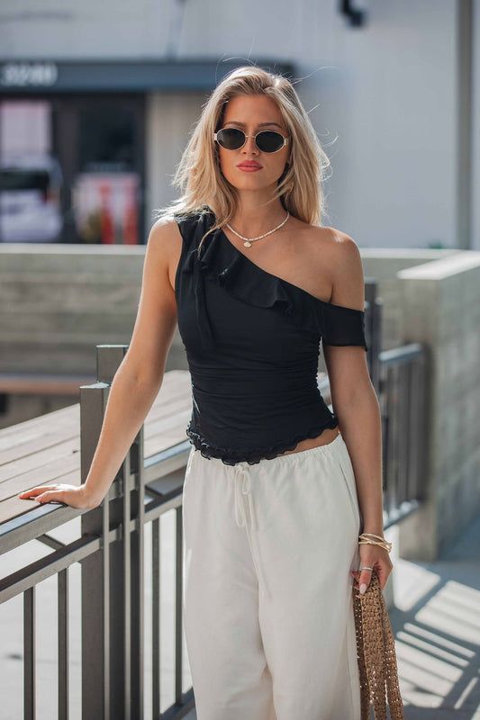 Woman in sunglasses, black Free People Isla Off The Shoulder Top, and white pants holds a woven bag by an outdoor railing.