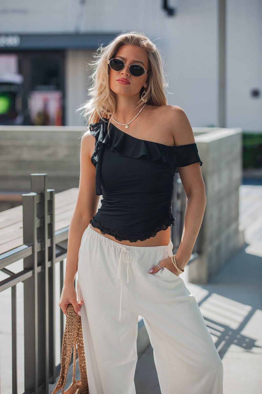 Woman in sunglasses wears the Free People Isla Off The Shoulder Top in black with white pants and a woven bag outdoors on a sunny day.