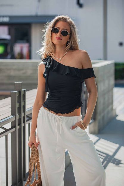 Woman in sunglasses wears the Free People Isla Off The Shoulder Top in black with white pants and a woven bag outdoors on a sunny day.