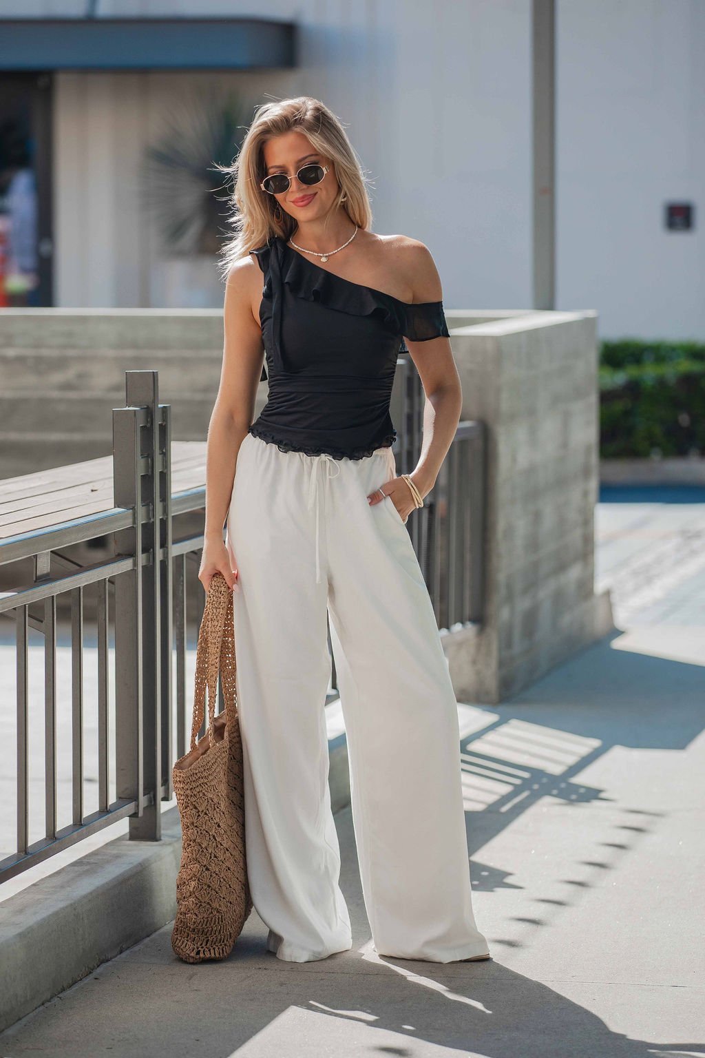 Woman in sunglasses wears the Free People Isla Off The Shoulder Top in black, white wide-leg pants, and a brown woven bag outdoors.