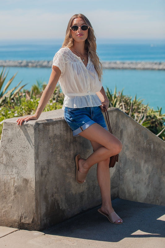 Woman in sunglasses, Free People Ivory Gemma Embroidered Top, and denim shorts leans on a concrete wall by the sea on a sunny day.