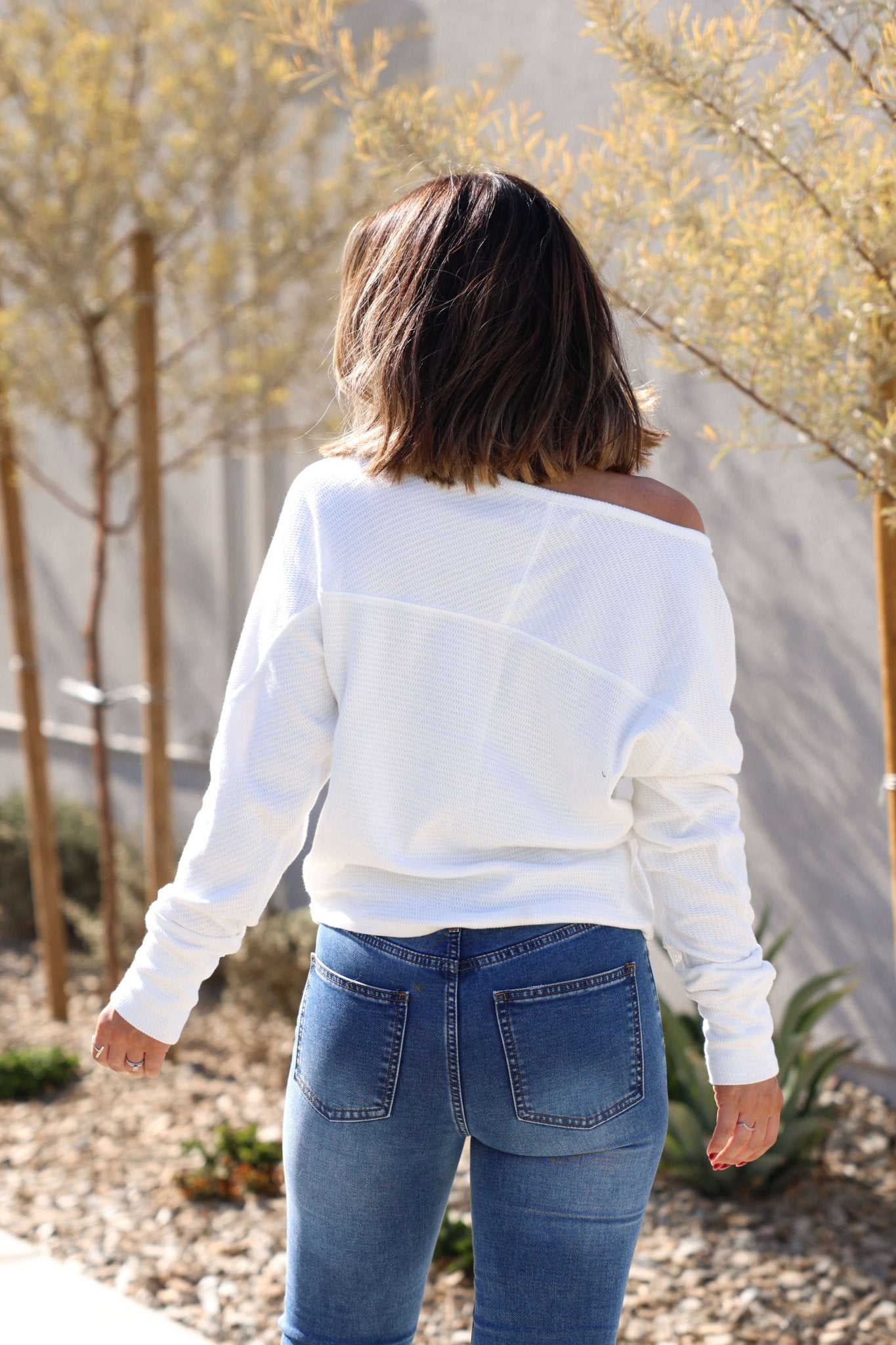 A woman in a Free People Ivory Jess Long Sleeve Top and blue jeans stands outdoors, facing away.