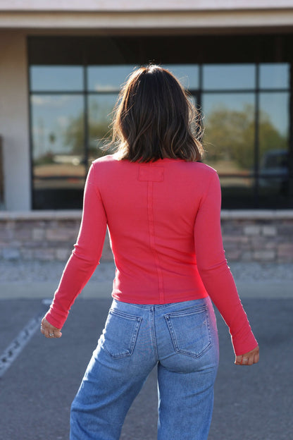 A woman in a Free People Light Red Neria Long Sleeve Top and blue jeans stands outdoors with her back to the camera.