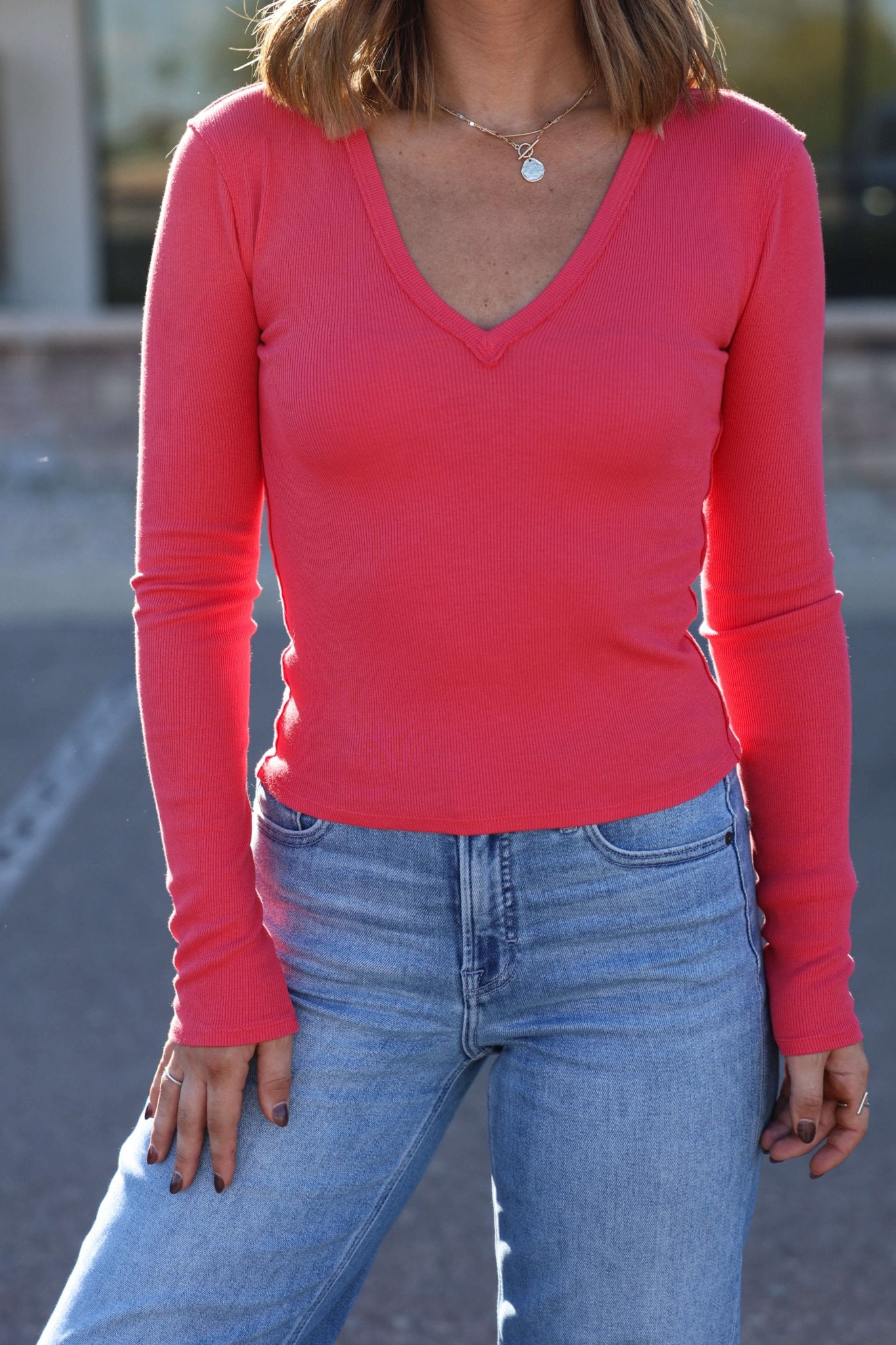 A woman in a Free People Light Red Neria Long Sleeve Top and light blue jeans stands outdoors, her face out of frame.