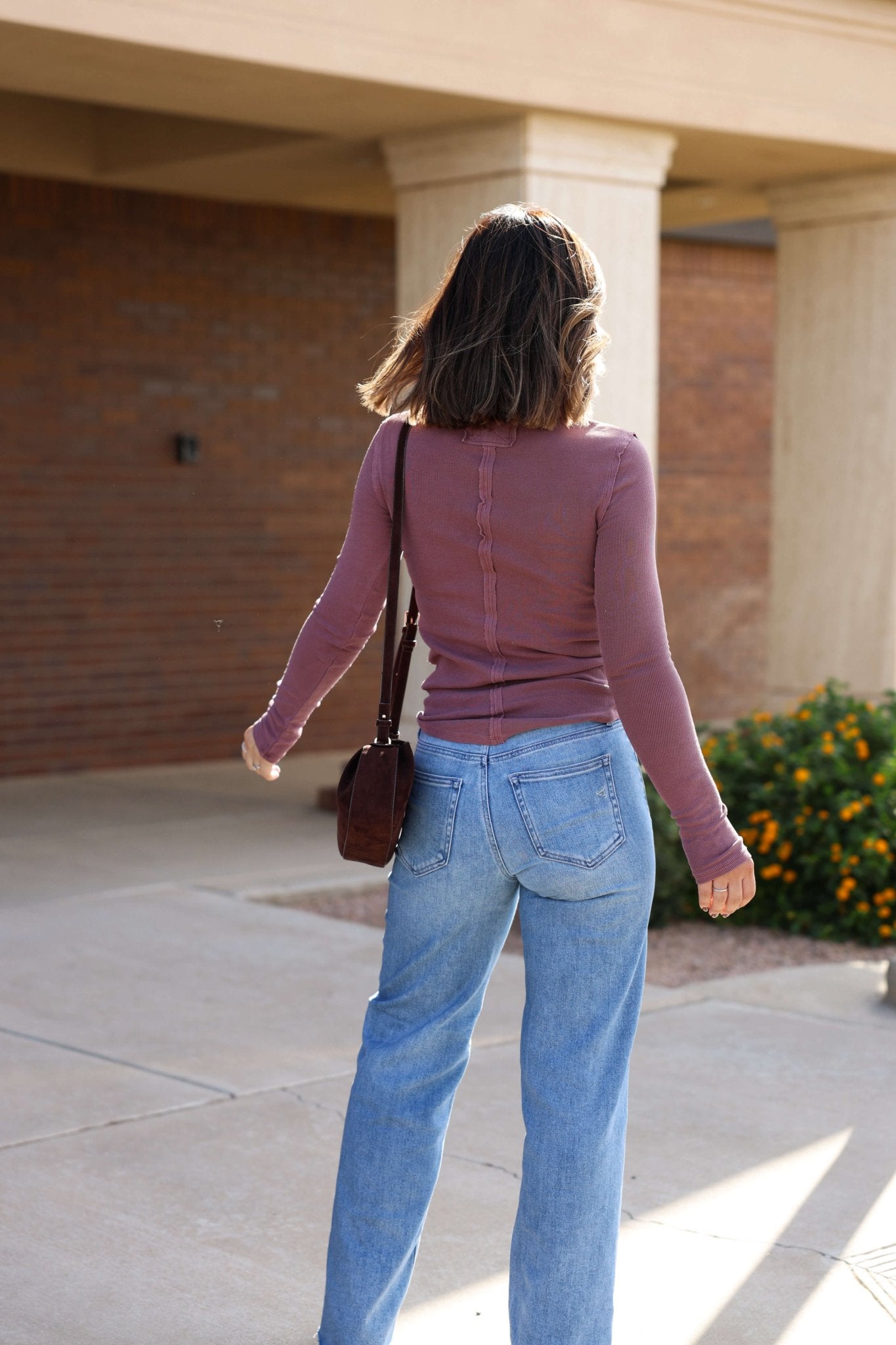 A woman in the Free People Mauve Jamie Henley Top and blue jeans stands outdoors, facing away.
