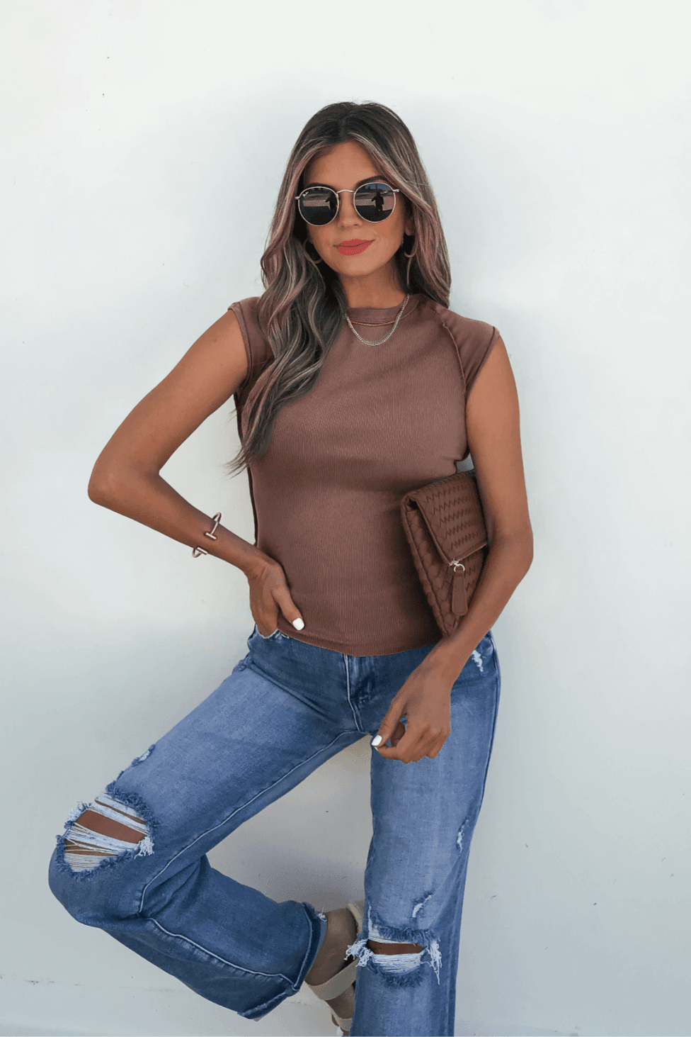 Woman in sunglasses wears the Free People Mocha Ever Had Tee and ripped jeans, holding a brown clutch against a white wall.