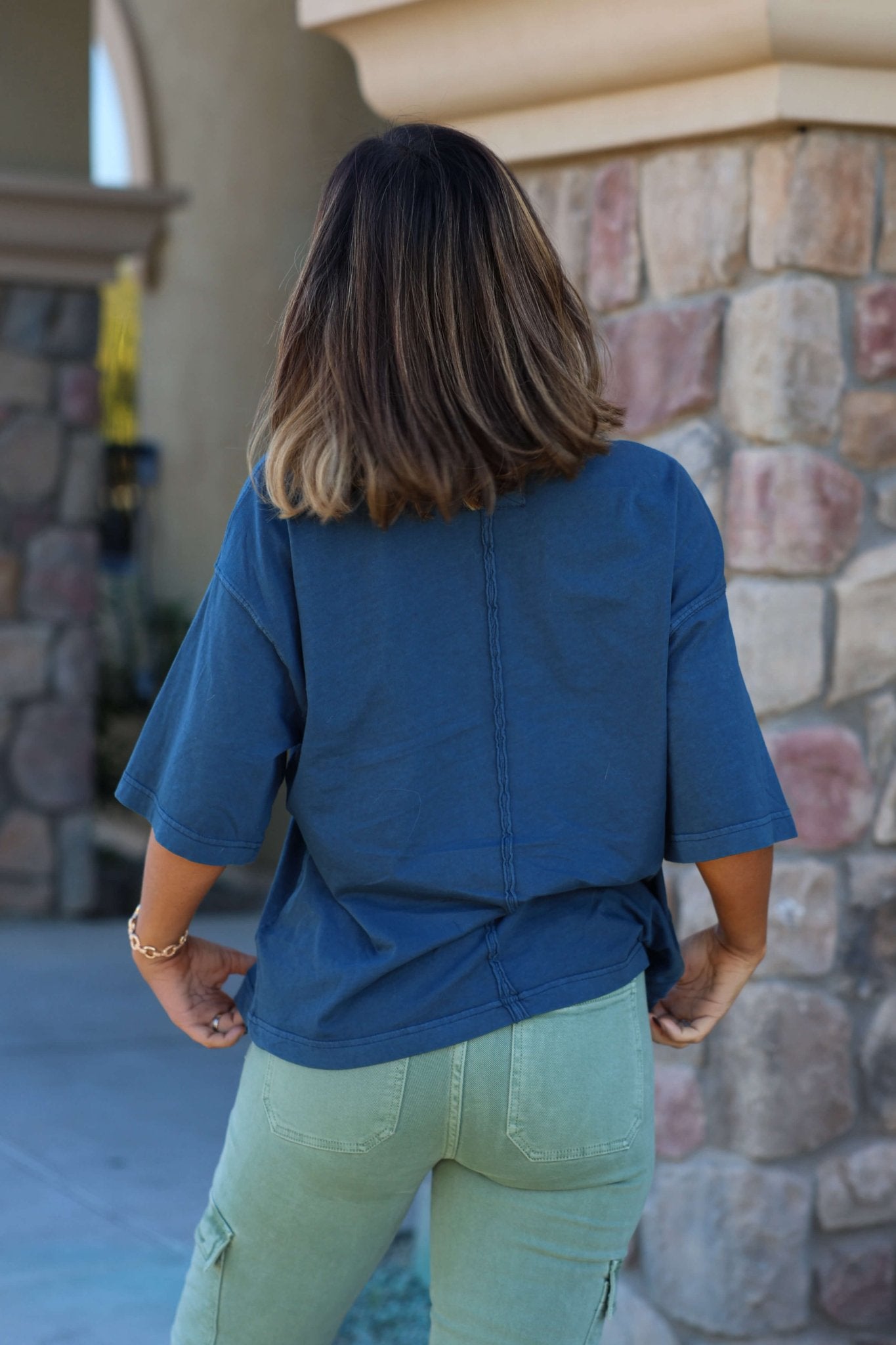 A person with shoulder-length brown hair wears a Free People Navy Perfect Oversized Tee and green pants by a stone wall, back facing camera.
