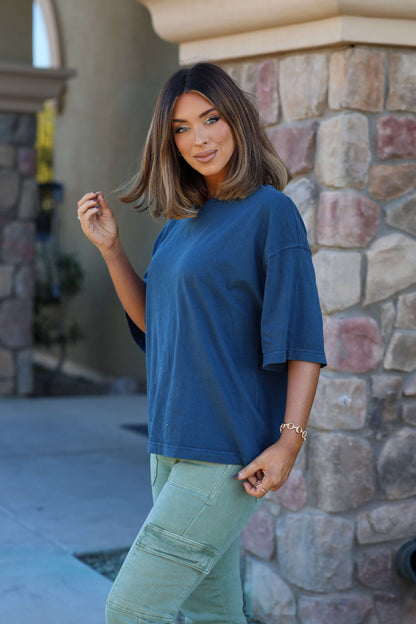 Woman with shoulder-length hair wears the Free People Navy Perfect Oversized Tee and green pants, smiling outdoors by a stone wall.