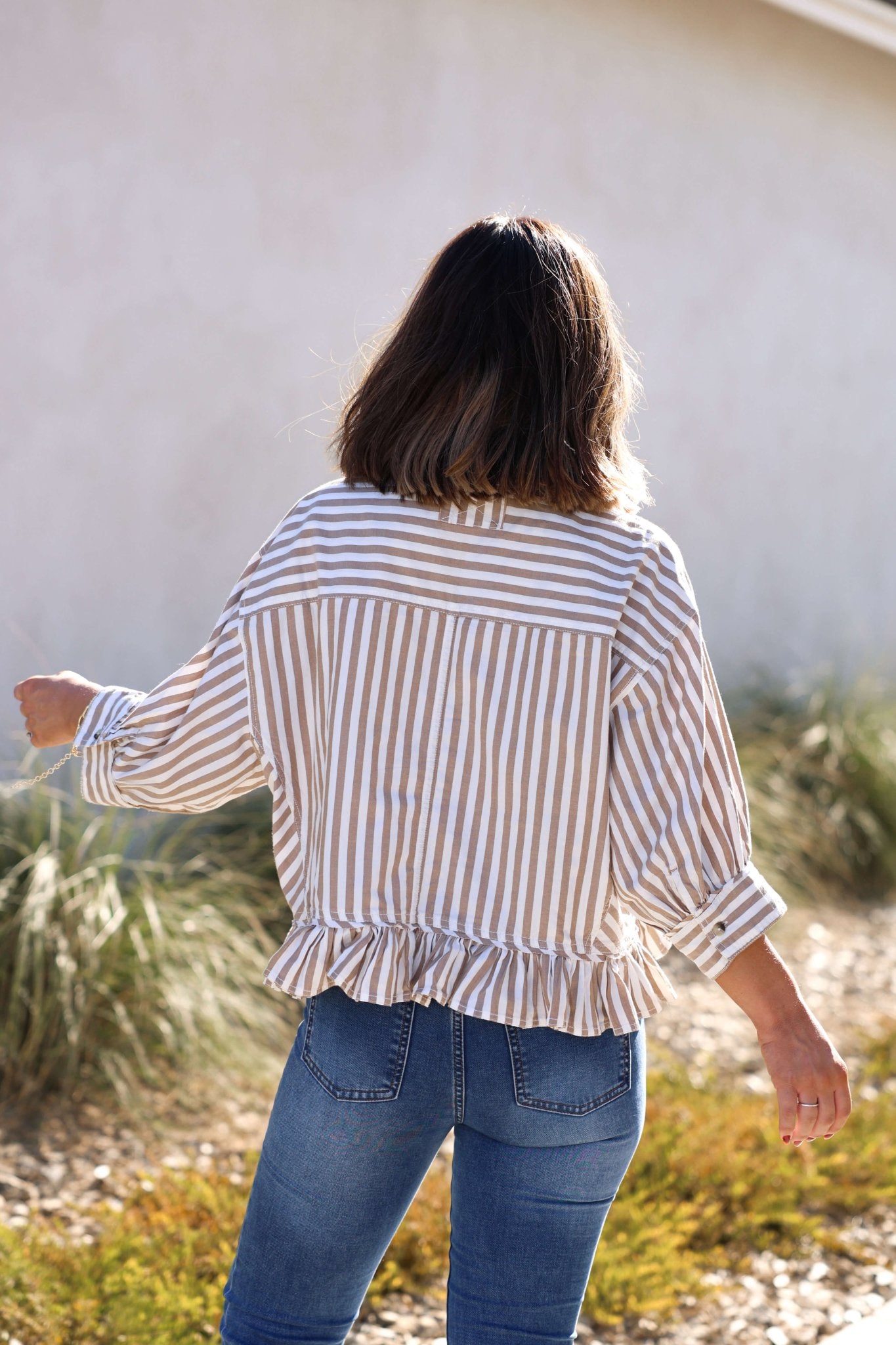 A woman with shoulder-length brown hair wears the Free People Neutral Bailey Stripe Shirt, standing outdoors with her back to the camera.