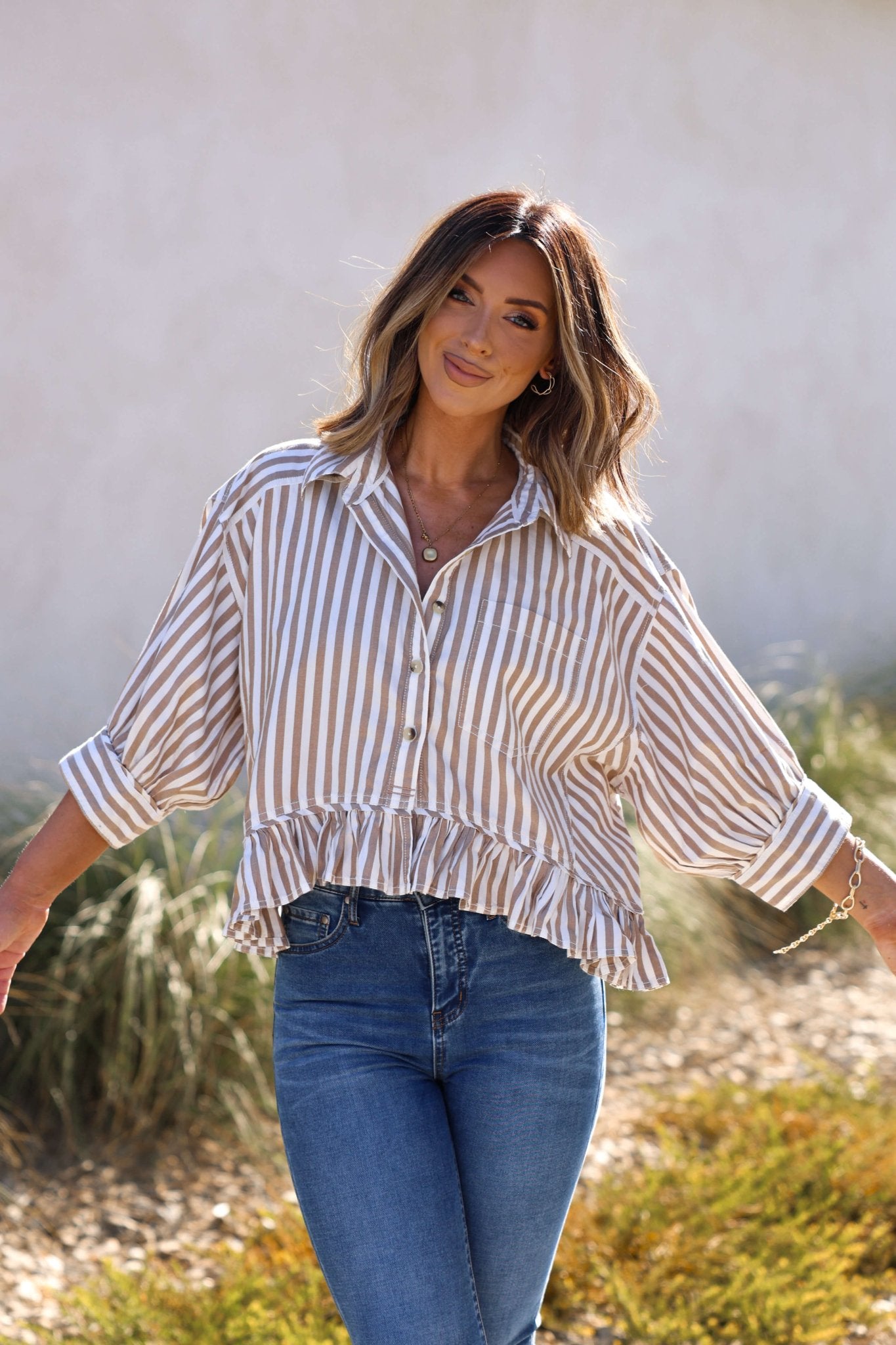 Woman smiling outdoors in the Free People Neutral Bailey Stripe Shirt – FINAL SALE, paired with blue jeans, arms outstretched.