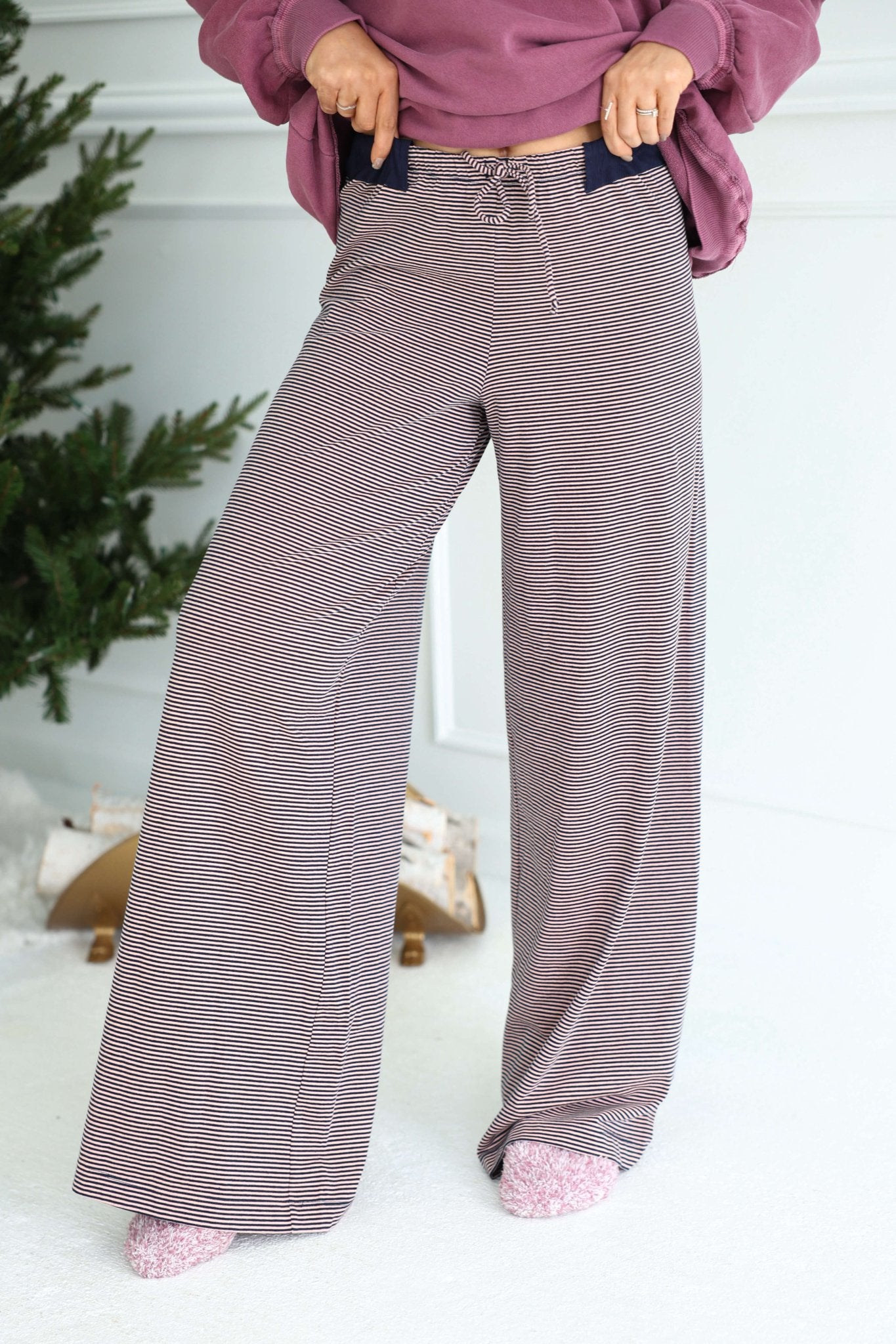 Person in Free People Peach Combo Out and About Pants, pink socks, and mauve sweatshirt stands by a Christmas tree indoors.