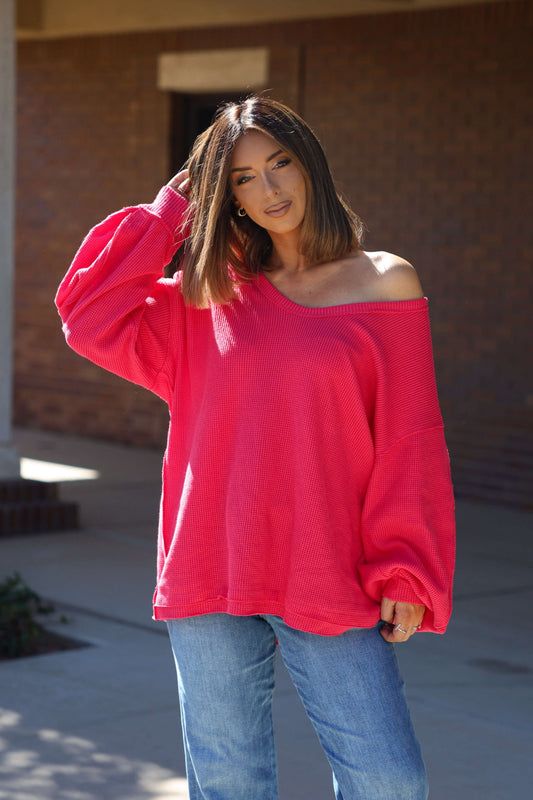A woman in a red Free People Coast 2 Coast Thermal Top and jeans stands outside, touching her hair and smiling slightly.