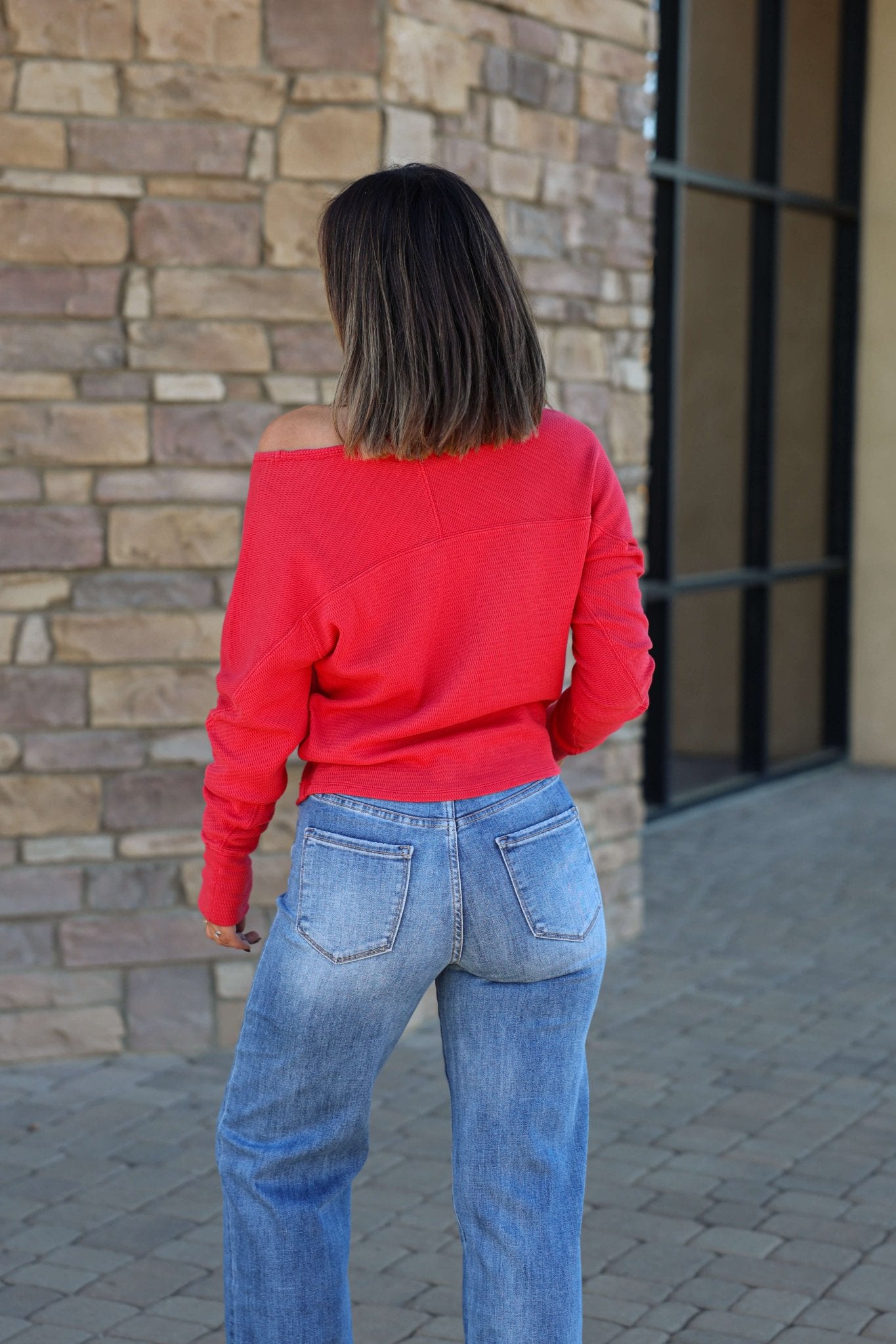 Wearing a Free People Red Jess Thermal Top and blue jeans, a person with shoulder-length hair stands on stone pavement by a brick wall.