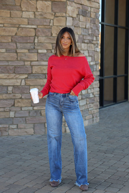 Woman in a Free People Red Jess Thermal Top and blue jeans holding coffee, standing on a stone path by a brick wall.