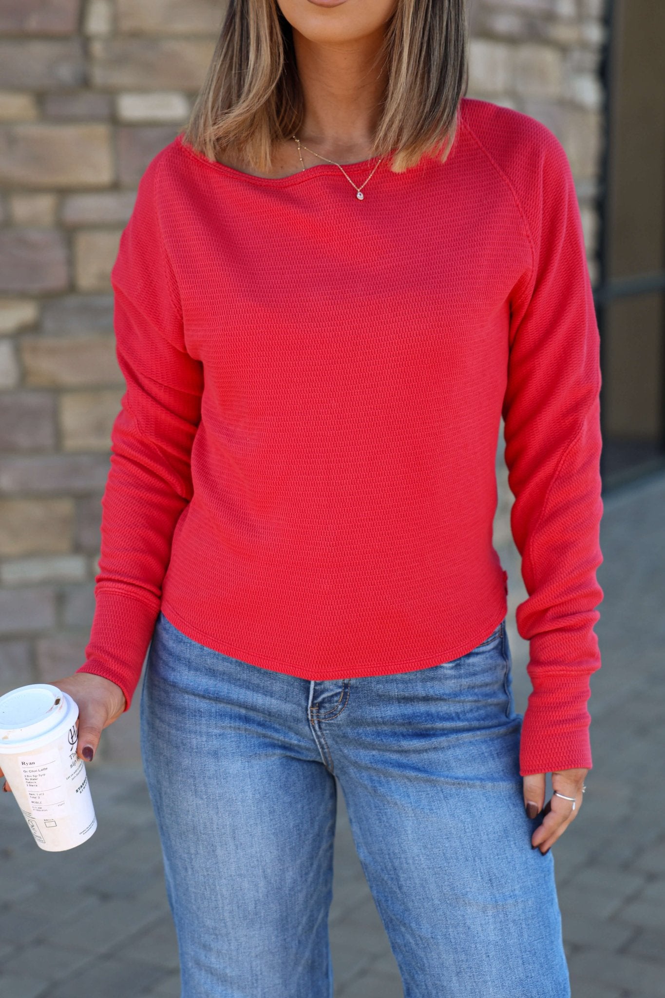 Woman wearing a Free People Red Jess Thermal Top and blue jeans holds a coffee cup in front of a brick wall.