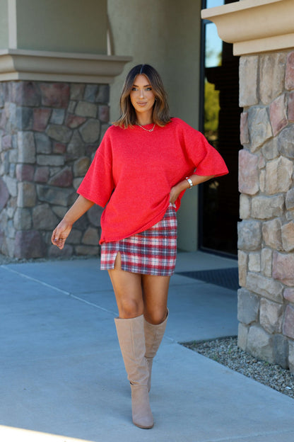 Woman wearing a Free People Red Margot Sweater Tee, plaid slit skirt, and tan knee-high boots poses outside by a stone building.