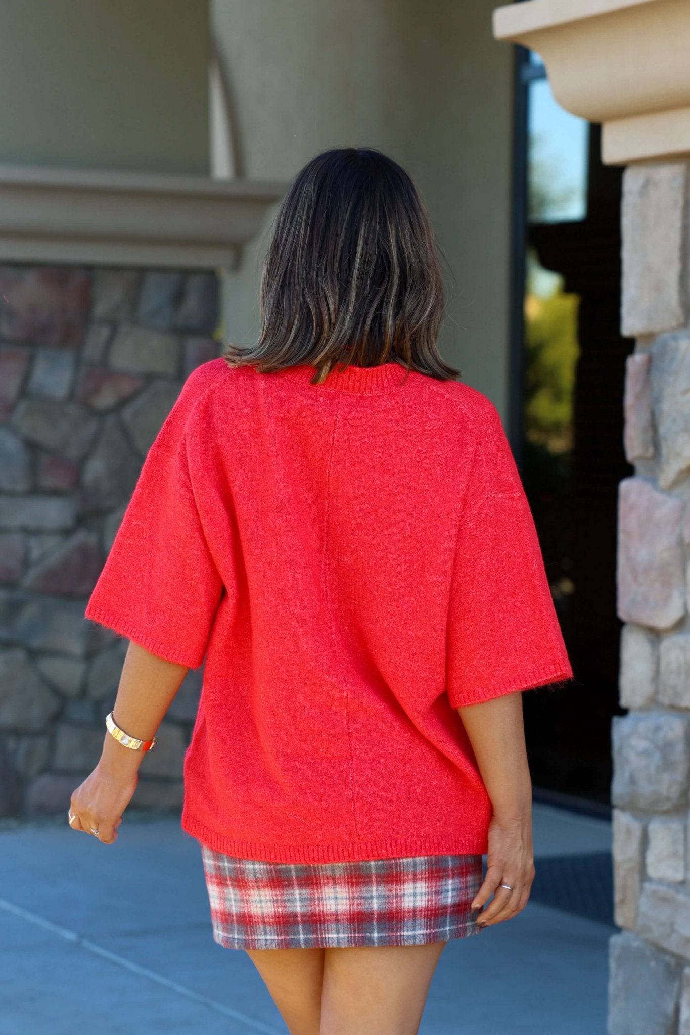 A woman in a Free People Red Margot Sweater Tee and plaid skirt stands outside, back to camera, near a stone building.
