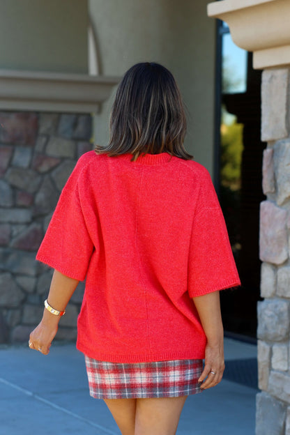 A woman in a Free People Red Margot Sweater Tee and plaid skirt stands outside, back to camera, near a stone building.