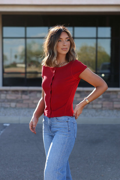 Wearing a Free People Red Most Wanted Top and blue jeans, a woman poses outside by a building with large windows.
