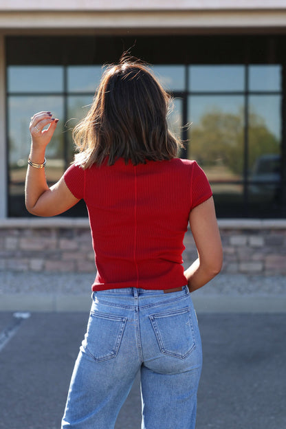 A woman in a Free People Red Most Wanted Top and blue jeans stands outside, facing away with her hand raised.