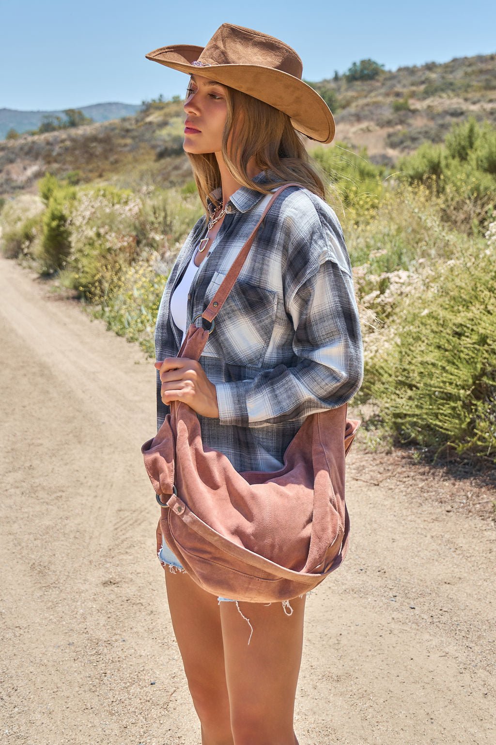 Woman in a plaid shirt, cowboy hat, and denim shorts carries a Free People Rust Roma Suede Tote Bag on a rural dirt road.