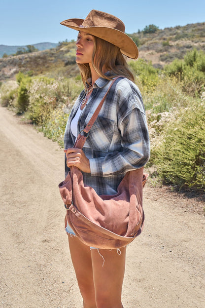 Woman in a plaid shirt, cowboy hat, and denim shorts carries a Free People Rust Roma Suede Tote Bag on a rural dirt road.