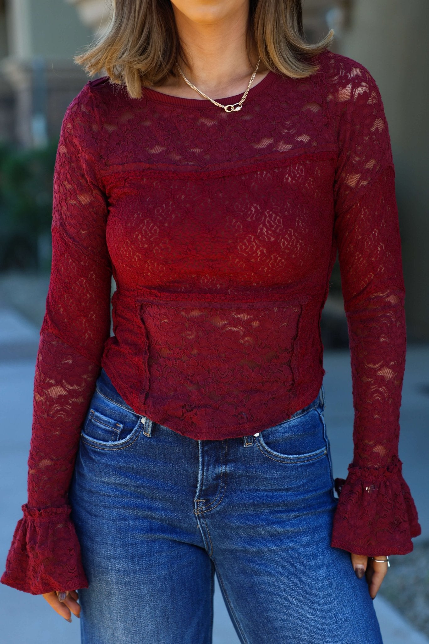 A woman wears the Free People Sheer Lace Flared Long Sleeve Top and blue jeans, standing outside with her hands by her sides.