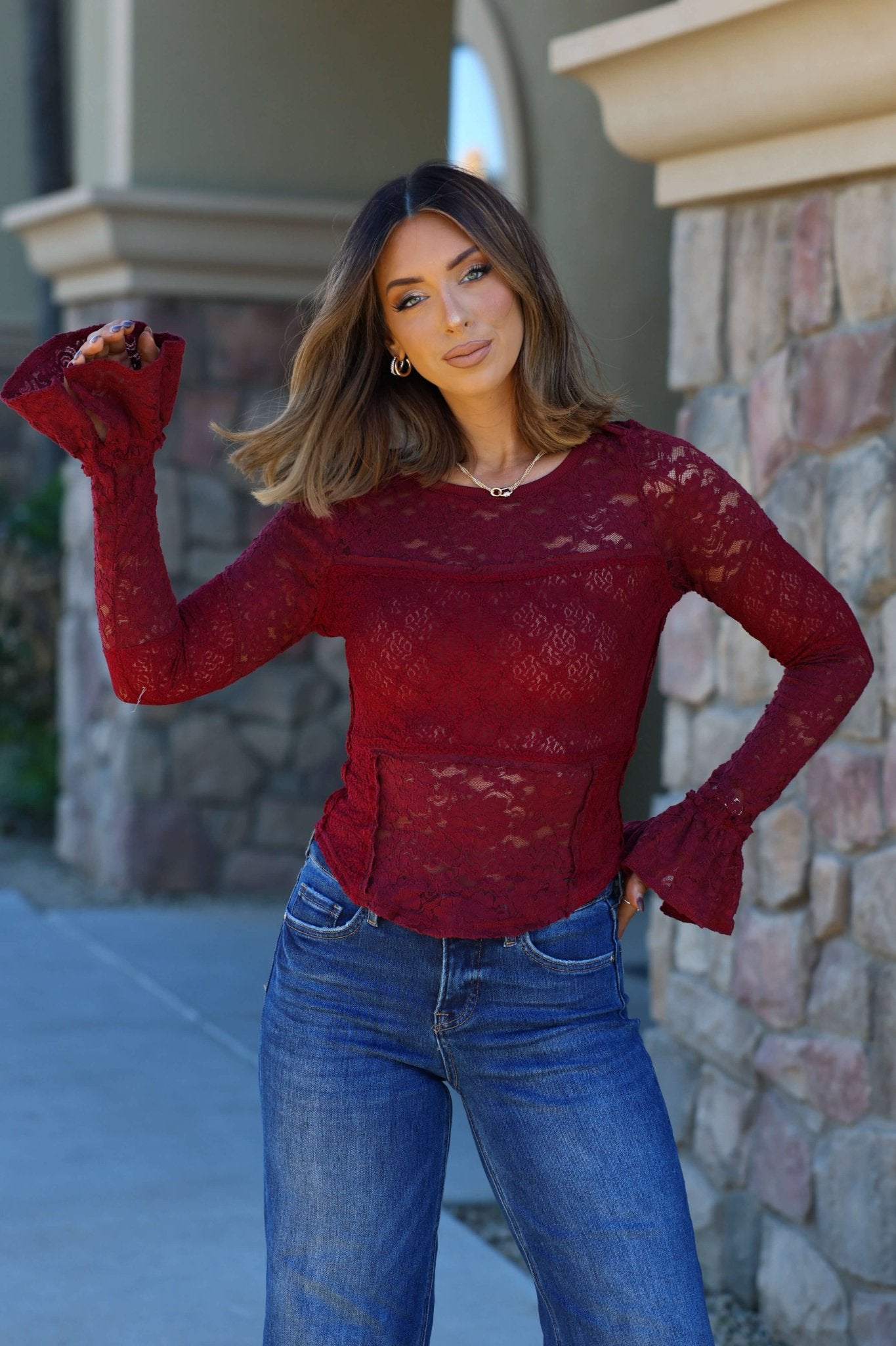 A woman poses confidently outdoors by a stone wall in a Free People Sheer Lace Flared Long Sleeve Top and blue jeans.