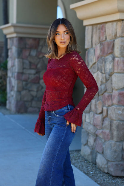 Woman in a Free People Sheer Lace Flared Long Sleeve Top and blue jeans stands by a stone wall, smiling to the side.