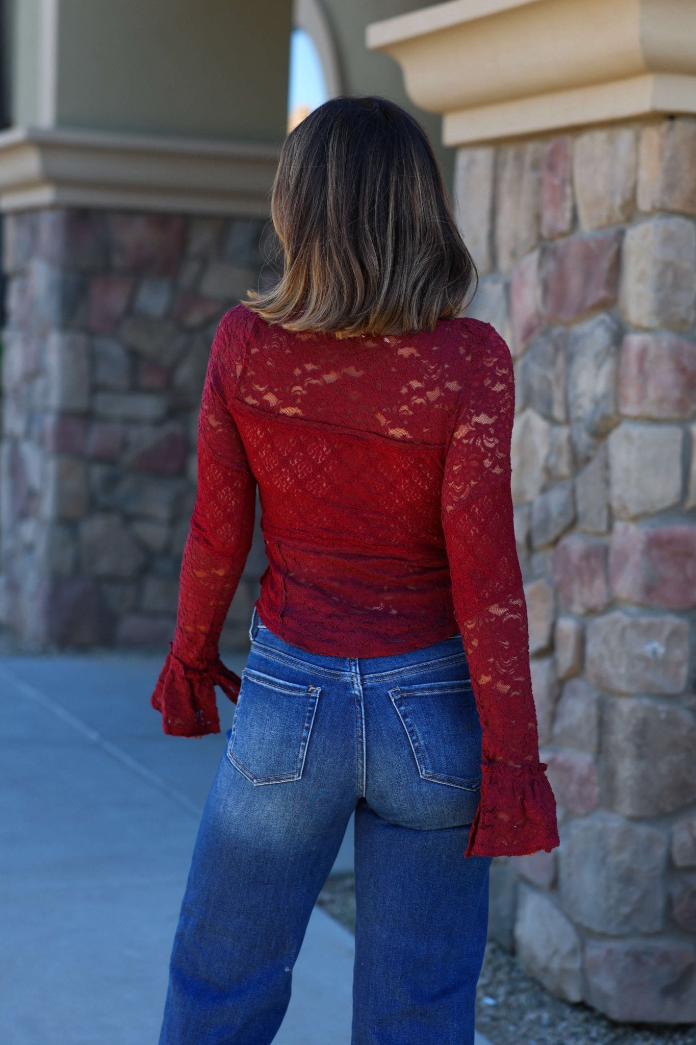 A woman wears a Free People Sheer Lace Flared Long Sleeve Top and blue jeans, standing outdoors by a stone wall.