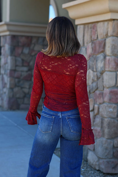 A woman wears a Free People Sheer Lace Flared Long Sleeve Top and blue jeans, standing outdoors by a stone wall.