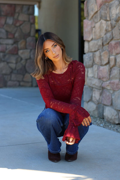 A woman in a Free People Sheer Lace Flared Long Sleeve Top and blue jeans squats on a sidewalk, looking at the camera.