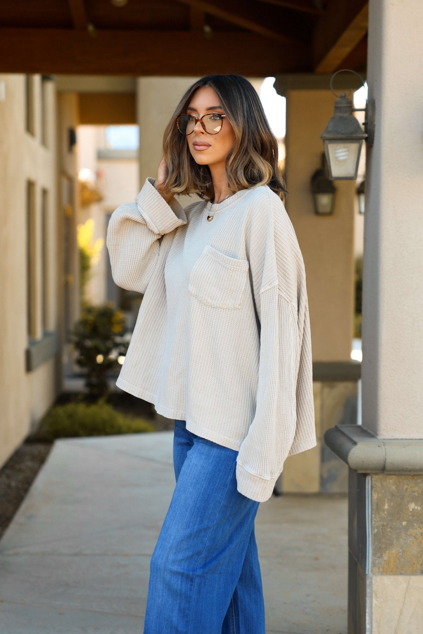 A woman in glasses wears a Free People Stone Dawson Pocket Thermal Top and blue jeans outdoors near a building.